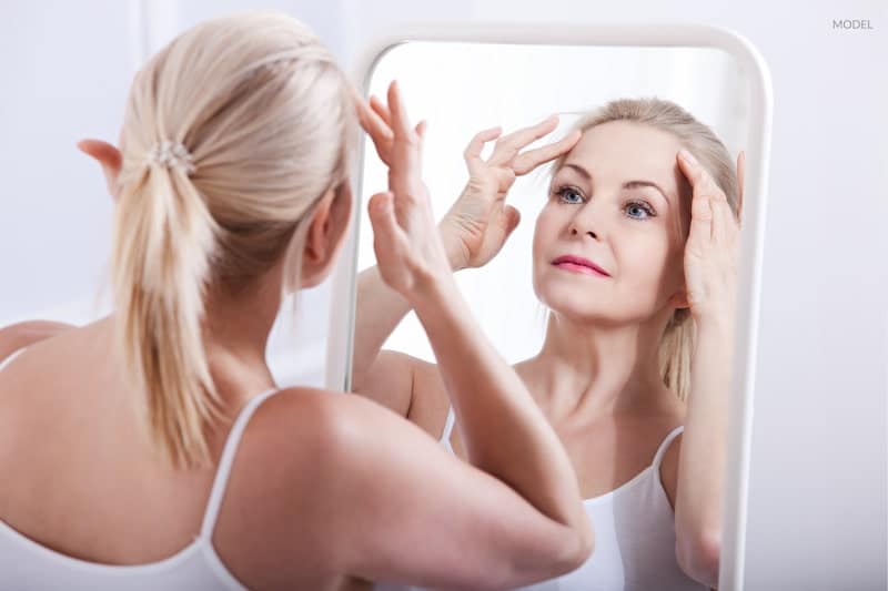 A woman examining her aging face in mirror, contemplating facelift surgery.