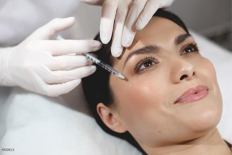 Women getting an injectable filler in the top of her cheek area by a woman in white surgical gloves.