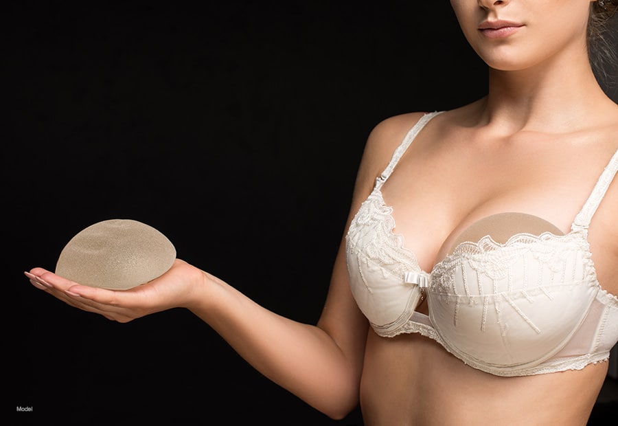A woman wearing a bra with a breast implant tucked inside holds another breast implant in her hand.